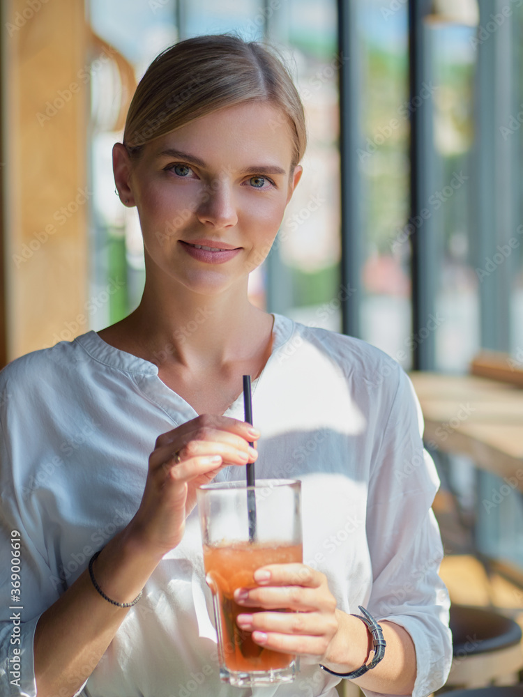 Calm beautiful happy young woman in relaxed mood looking outdoors and smiling sitting at the bar counter near big window with healthy delicious fresh lemonade glass dressed trendy white shirt Calm beautiful happy young woman in relaxed mood looking outdoors and smiling sitting at the bar counter near big window with healthy delicious fresh lemonade glass dressed trendy white shirt