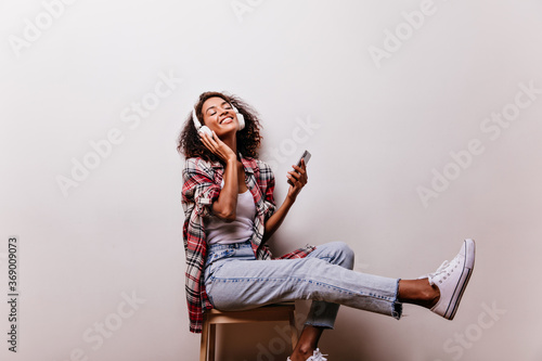 Stunning black girl in jeans sitting in headphones on white background. Chilling african lady in checkered shirt listening music.