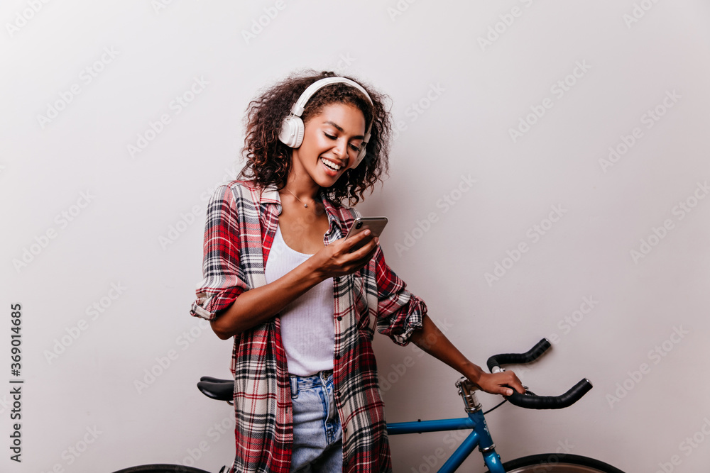 Enchanting stylish woman in headphones reading phone message. Well-dressed african girl with bicycle holding smartphone. Enchanting stylish woman in headphones reading phone message. Well-dressed african girl with bicycle holding smartphone.