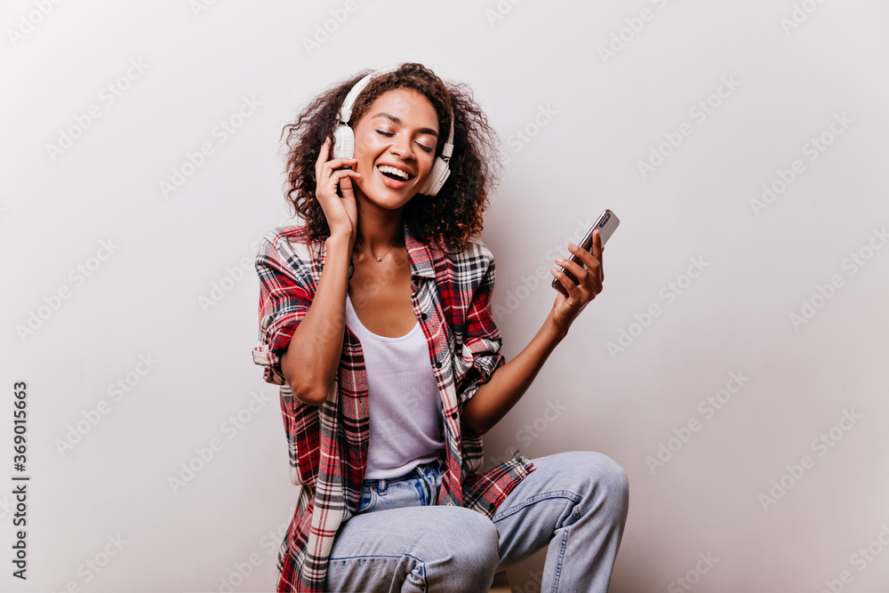 Happy black girl in red shirt listening music in earphones. Blithesome young woman with curly hairstyle chilling with favorite song.