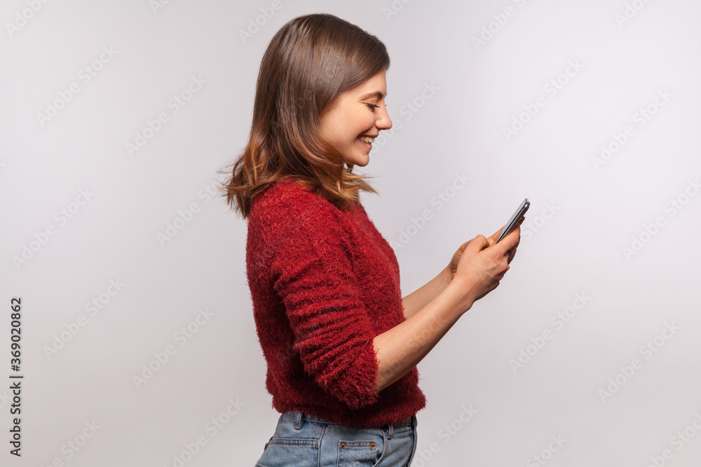 Side view, happy brunette girl in shaggy sweater using mobile phone and smiling laughing joyfully, typing or reading funny message on social network, online communication. studio shot isolated on gray Side view, happy brunette girl in shaggy sweater using mobile phone and smiling laughing joyfully, typing or reading funny message on social network, online communication. studio shot isolated on gray