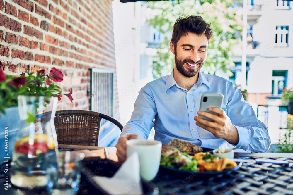 Cheerful young entrepreneur eating breakfast in outdoor cafe and using smartphone