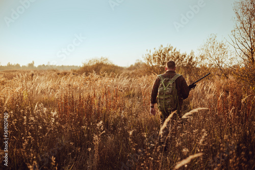 Young hunter in the sunny field with military backpack and shotgun waiting for flying ducks
