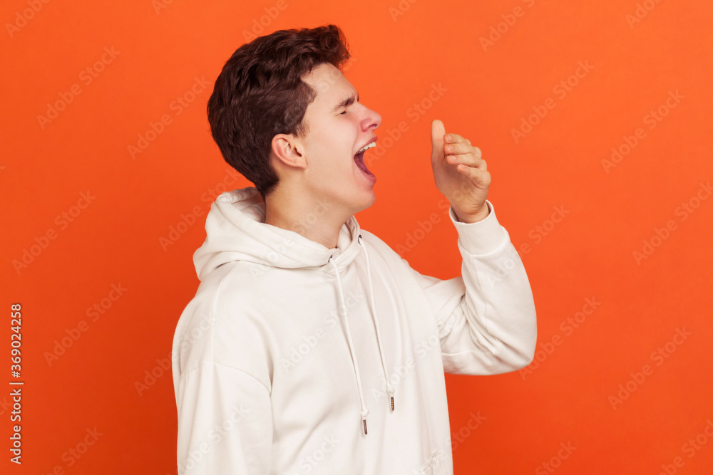 Profile of bored young man in casual style sweatshirt with hood yawning, sleepy teenager exhausted of early awakenings, laziness. Indoor studio shot isolated on orange background Profile of bored young man in casual style sweatshirt with hood yawning, sleepy teenager exhausted of early awakenings, laziness. Indoor studio shot isolated on orange background