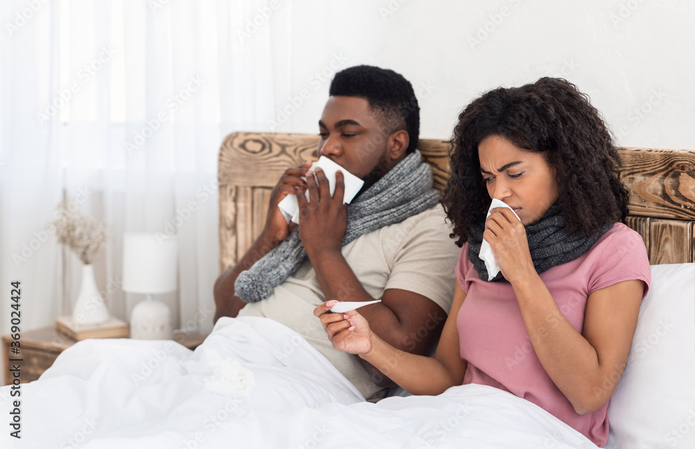 Sick black couple sitting in bed, measuring fever, sneezing noses Sick black couple sitting in bed, measuring fever, sneezing noses