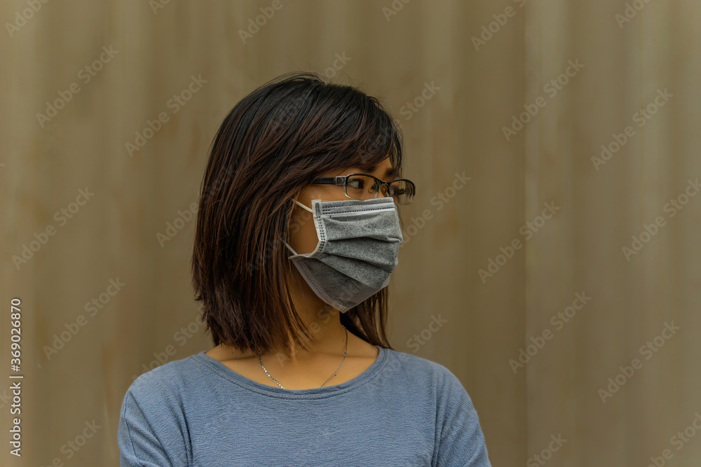 Asian young attractive woman wearing a protective hygiene mask over her face to protect flu and virus. Unwell influenza in infected woman portrait with light brown background Asian young attractive woman wearing a protective hygiene mask over her face to protect flu and virus. Unwell influenza in infected woman portrait with light brown background