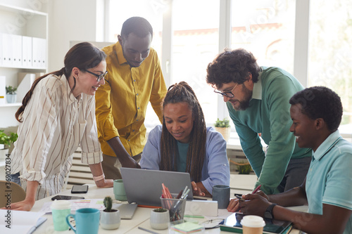 Portrait of contemporary multi-ethnic team leaning over laptop and smiling while working together in office, copy space photo