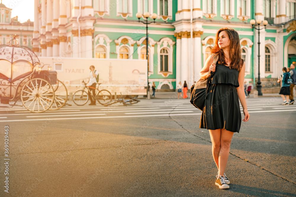 A young brunette woman in a dress with long hair with a backpack stands at full height against the background of the winter Palace on the Palace square in St. Petersburg