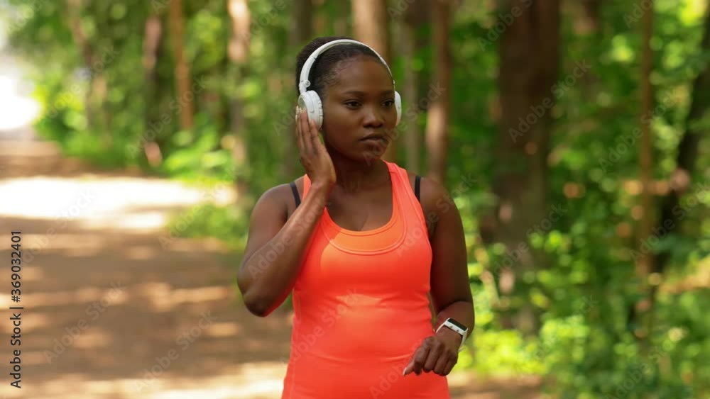 fitness, sport and technology concept - young african american woman with headphones listening to music on smart watch running in forest fitness, sport and technology concept - young african american woman with headphones listening to music on smart watch running in forest
