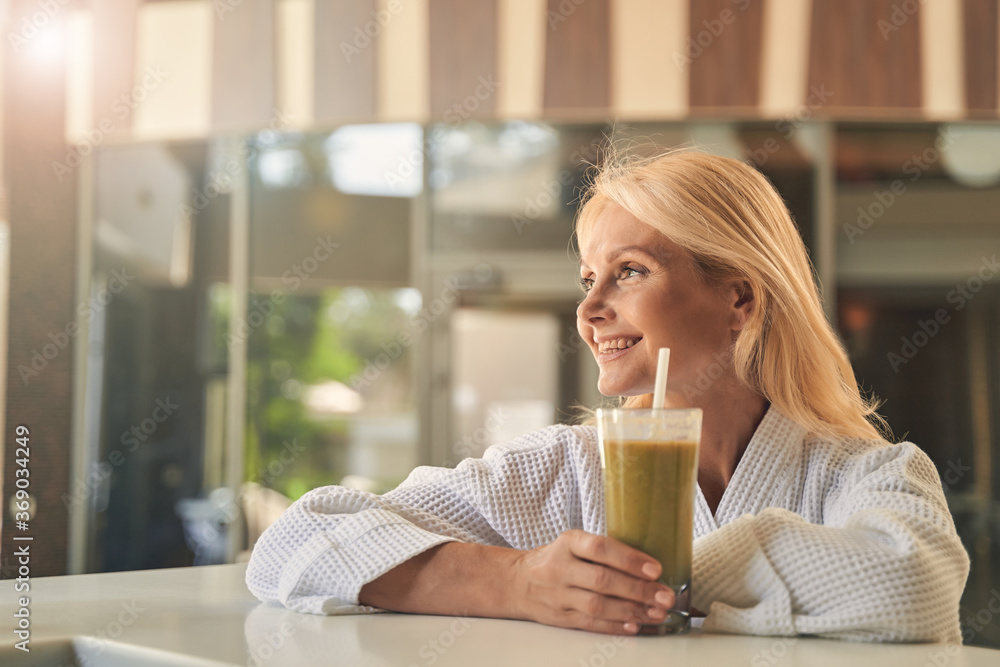 Cheerful woman in white bathrobe holding glass of juice while looking away Cheerful woman in white bathrobe holding glass of juice while looking away