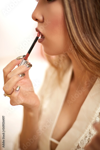 a young beautiful Asian woman in a beige lace dressing gown put on morning makeup and puts lipstick on her lips. in the bedroom . shallow depth of focus. selective focus