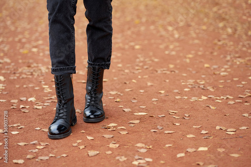 Woman's legs in fashionable autumn leather black boots and trendy black jeans. Trendy youth casual outfit for everyday. Modern street collection. Autumn road with yellow leaves on background.