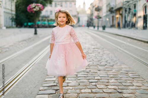 Cute blond little girl walks along the street of the old European city in her beautiful pink dress. Front view. Copy space © sofiko14