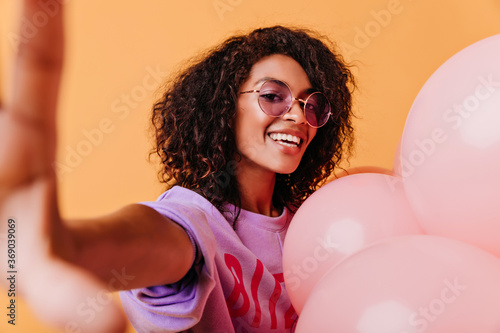 Blithesome girl with wavy hair posing with excitement on orange background. Jocund african woman making selfie with helium balloons. © Look!