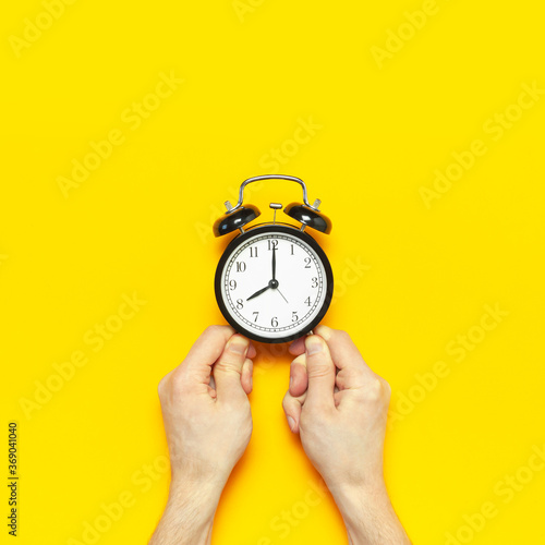 Black vintage alarm clock in male hands on yellow background flat lay copy space. Retro clock, mechanical alarm clock. Minimalistic background, concept of time, deadline, time to work, morning