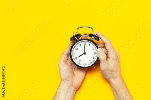 Black vintage alarm clock in male hands on yellow background flat lay copy space. Retro clock, mechanical alarm clock. Minimalistic background, concept of time, deadline, time to work, morning