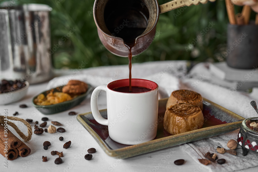 Coffee preparation. pouring coffee from copper pot into a cup on a white table with cinnamon buns