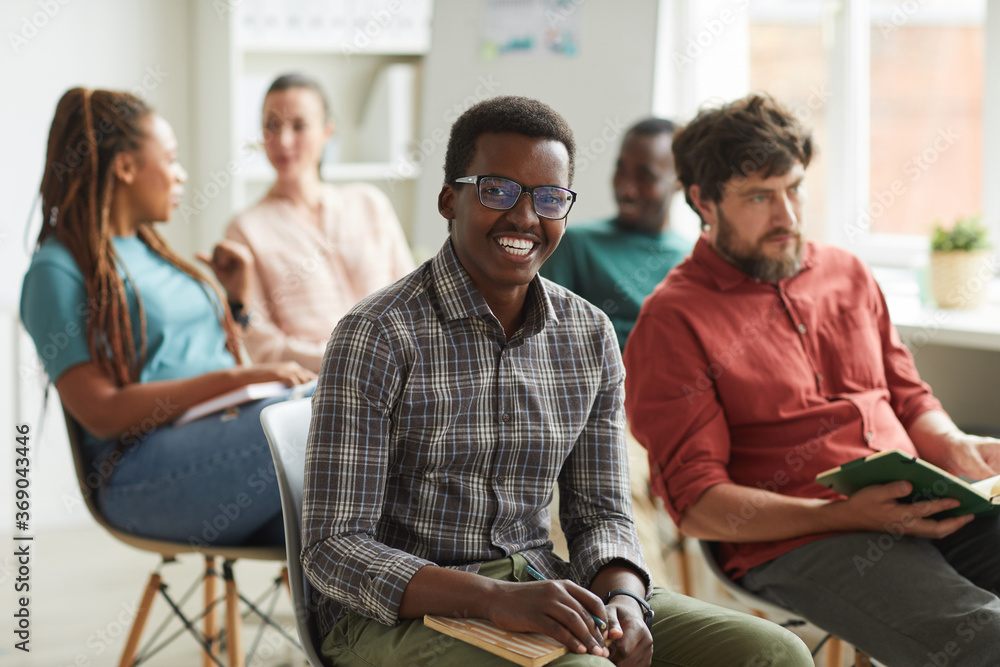 Obraz premium Multi-ethnic group of people sitting in audience during training seminar or business conference in office, focus on young African-American man smiling at camera in foreground, copy space
