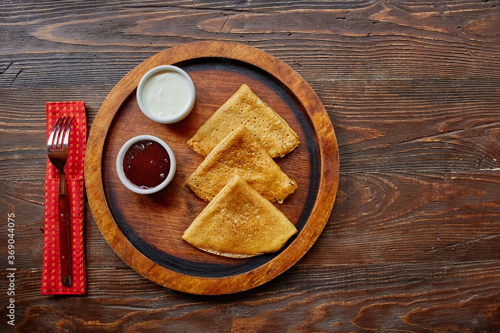 Homemade hot delicious pancakes with sour cream and jam on a wooden plate, top view. Homemade hot delicious pancakes with sour cream and jam on a wooden plate, top view.