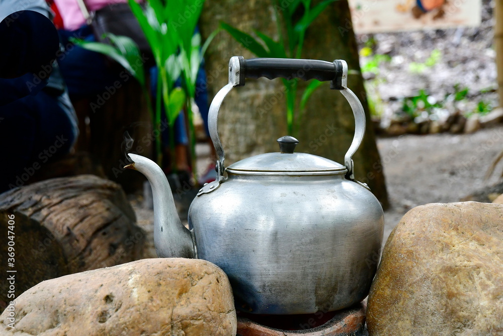 A large antique kettle made of aluminum, placed on a charcoal stove beside the stone. Boiling water pot is used for boiling hot water to prepare hot beverages such as tea, coffee or warm water. 

