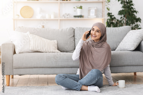 Cheerful arabic girl in headscarf talking on cellphone while relaxing at home