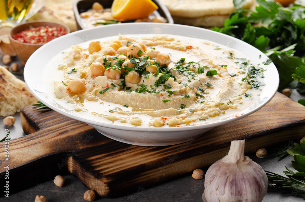 Fresh homemade Hummus in clay dish topped with olive oil, chickpeas and chopped green coriander leaves on stone table served with spices