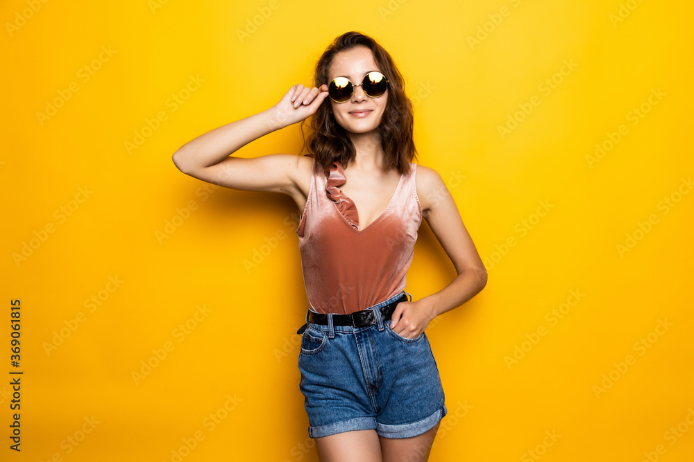 Young happy girl in summer hat wearing sunglasses in summer clothes smiling on yellow background