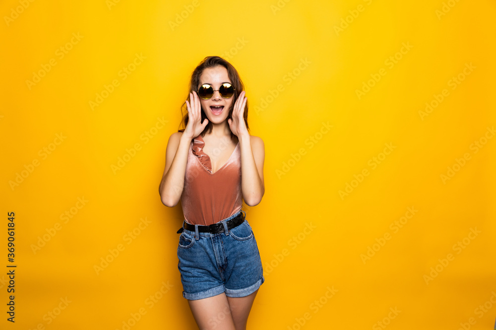 Young woman sunglasses looking away with surprised smile isolated on yellow background.