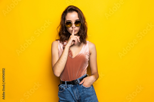 Portrait of a pretty young girl showing silence gesture and winking isolated over yellow background