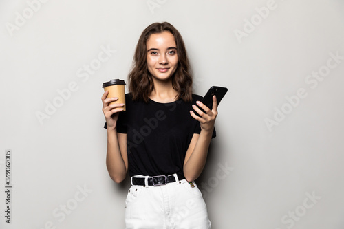 Portrait of a young smiling girl in shirt texting message on mobile phone and holding cup of coffee to go over gray background