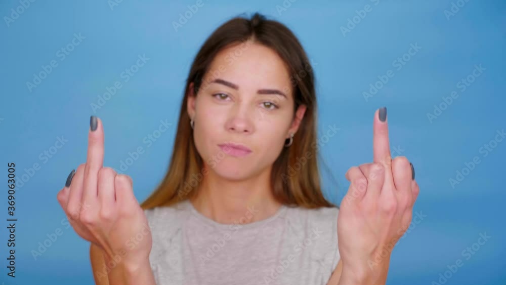 Positive woman in gray t-shirt smiles, raises hands and shows middle finger on blue background with copy space. 4K slow motion footage Positive woman in gray t-shirt smiles, raises hands and shows middle finger on blue background with copy space. 4K slow motion footage