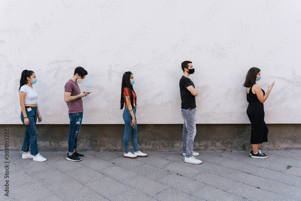 Fototapeta premium Group of young people at safety distance near a white wall in queue in protection by Coronavirus, Covid-19 with face mask while using the device - Multiracial group of friends - Concept of security