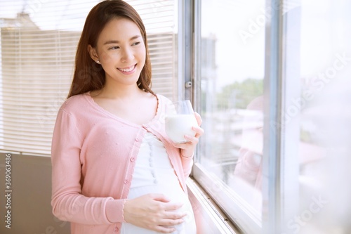 Beautiful pregnant woman standing by the window with a glass of milk in her hand