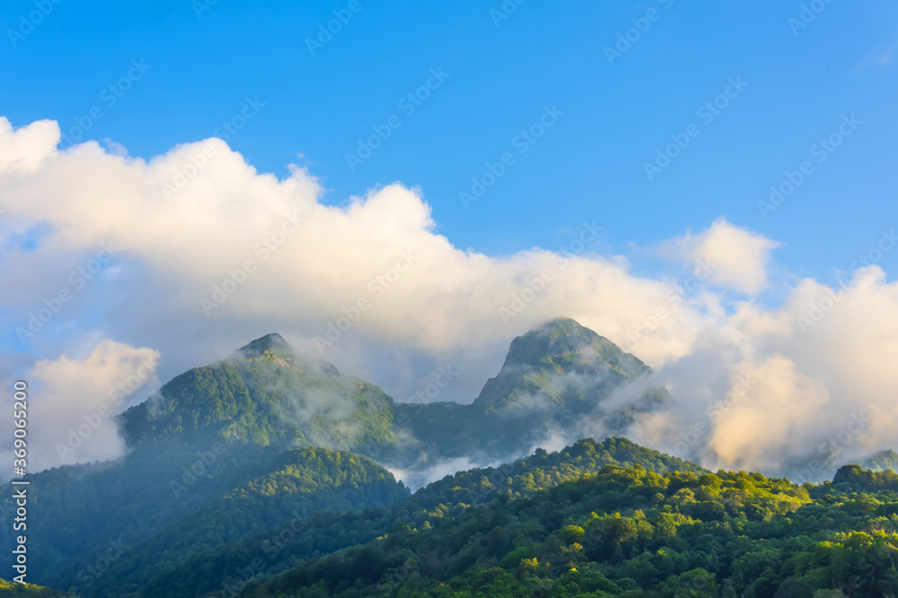View of two mountain peaks with clouds and covered with forests at the foot, in the evening at sunset light. View of two mountain peaks with clouds and covered with forests at the foot, in the evening at sunset light.