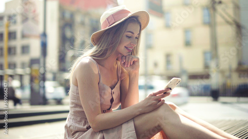 Young blonde woman wearing hat using phone in a city.