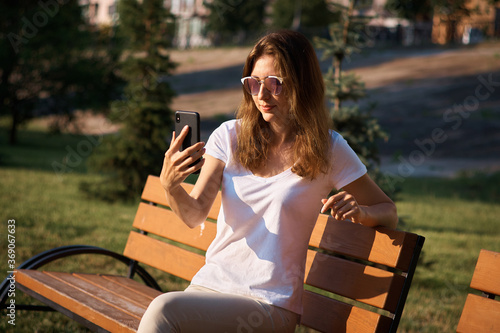 Girl blogger writes a post in the park. A young girl with a phone in her hands sits on a bench. ?ommunication via phone. chatting in the park photo