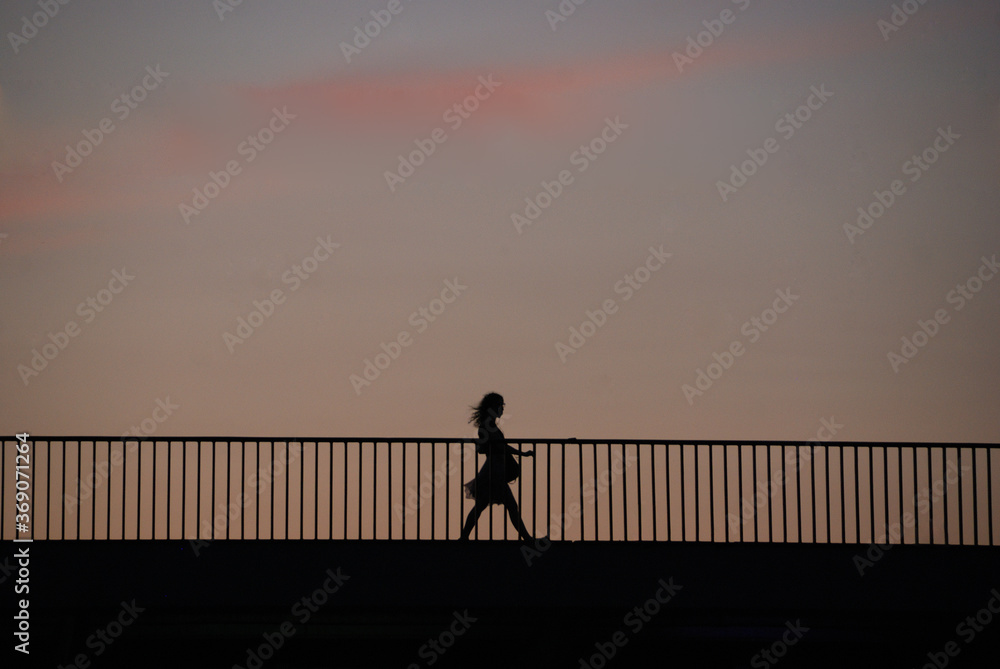Silhouette of a pregnant woman in a flowy dress crossing a bridge at sunset