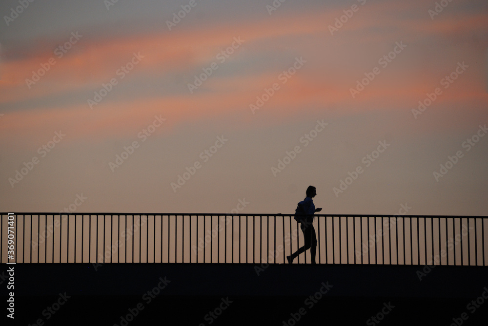 Silhouette of a woman with a backpack and a cell phone in her hand crossing a bridge at sunset