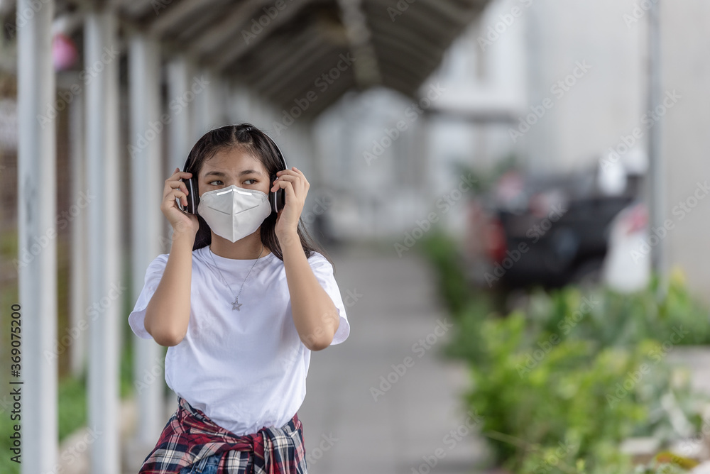 Asian young woman walking on street with her headphones while wearing face mask as protection against infectious diseases during coronavirus pandemic. Basic equipment to help keep us safe new life.