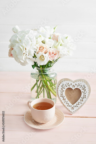 Bouquet of roses, eustomas and hydrangeas on table. Cup of tea and photo frame. Greeting card for mothers day. Cozy good morning. Happy Birthday. Romantic breakfast. Flowers composition for Valentine