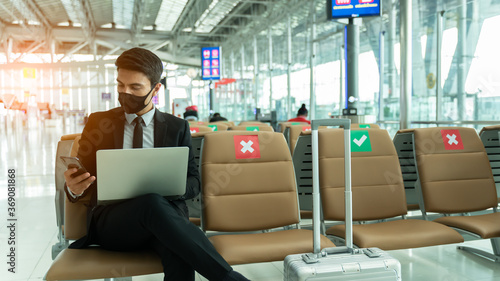 New normal and social distance concept.Business man wearing mask using smartphone searching airline flight status and sitting with distance during corona virus 2019 outbreak at airport