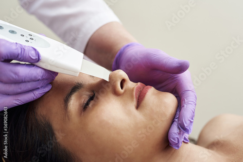 A woman makes an ultrasonic cleaning of the face and skin to a client. Modern equipment. Young pretty woman receiving treatments in beauty salons and lying on the couch