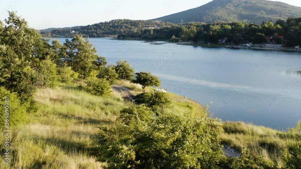 Flight with a drone from a grassy coast of a mountain lake surrounded by vacation homes and a thick forest with scenic background Flight with a drone from a grassy coast of a mountain lake surrounded by vacation homes and a thick forest with scenic background