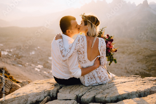 Beautiful bride and bridegroom kissing and hugs sitting on stone cliff at sunset