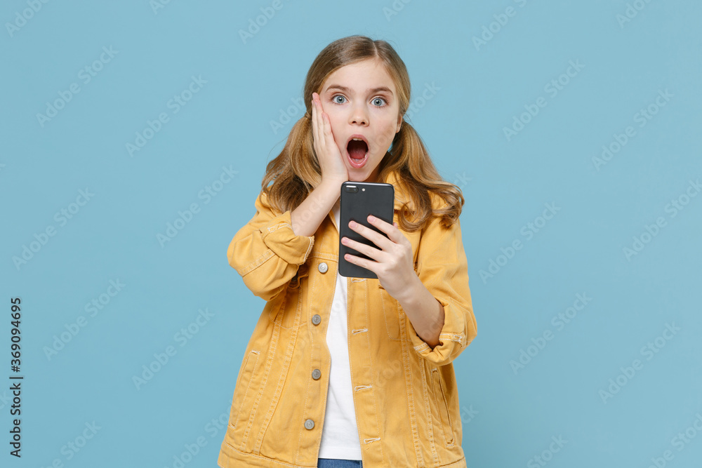 Shocked little blonde kid girl 12-13 years old in yellow jacket isolated on pastel blue wall background studio. Childhood lifestyle concept. Mock up copy space. Using mobile phone, put hand on cheek.
