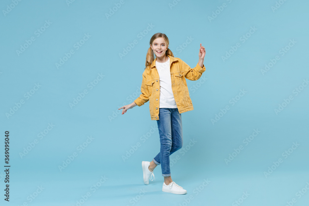 Full length portrait of funny little blonde kid girl 12-13 years old in yellow jacket isolated on blue background. Childhood lifestyle concept. Mock up copy space. Pointing index finger up, dancing. Full length portrait of funny little blonde kid girl 12-13 years old in yellow jacket isolated on blue background. Childhood lifestyle concept. Mock up copy space. Pointing index finger up, dancing.