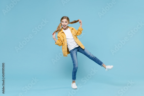 Full length portrait of excited little blonde kid girl 12-13 years old in yellow jacket posing isolated on blue wall background studio. Childhood lifestyle concept. Mock up copy space. Hold ponytails.