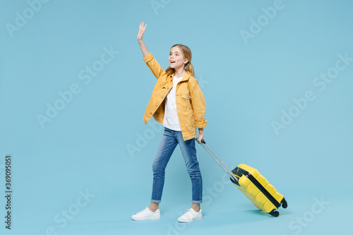 Full length portrait of little kid girl 12-13 years old isolated on blue background. Passenger traveling abroad on weekends. Air flight journey concept Hold suitcase waving greet with hand catch taxi.