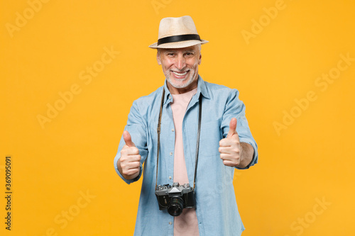Smiling traveler tourist elderly gray-haired man in hat with photo camera isolated on yellow background. Passenger traveling abroad on weekends getaway. Air flight journey concept. Showing thumbs up.