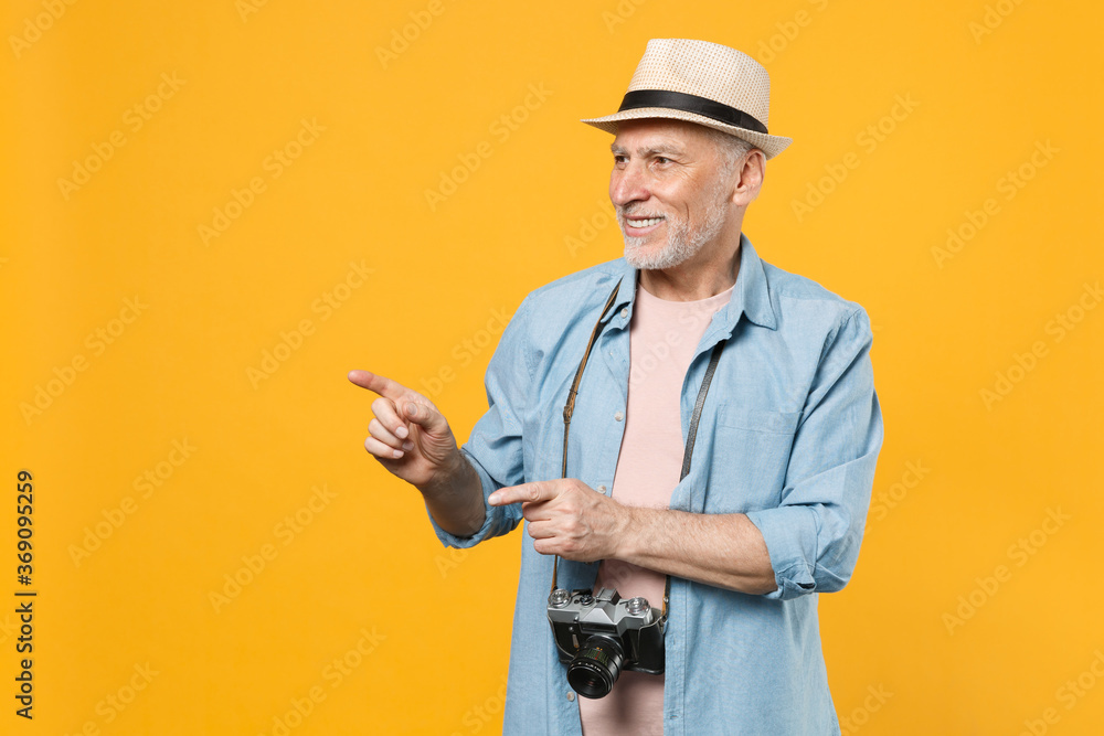 Smiling traveler tourist elderly gray-haired man in hat photo camera isolated on yellow background. Passenger traveling abroad on weekend getaway. Air flight journey concept Point index fingers aside. Smiling traveler tourist elderly gray-haired man in hat photo camera isolated on yellow background. Passenger traveling abroad on weekend getaway. Air flight journey concept Point index fingers aside.